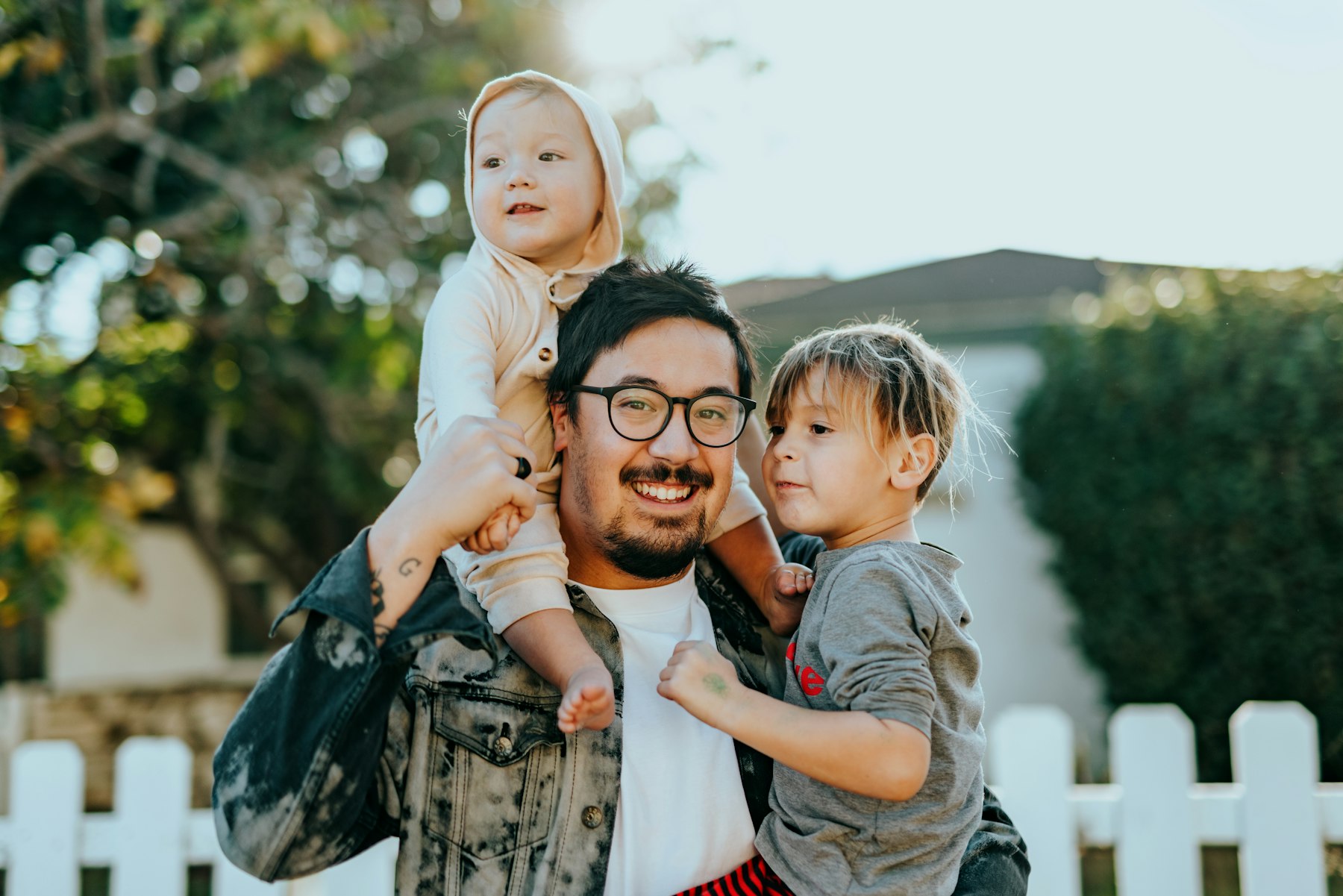 A dad laughing with his two young kids on his shoulders outside at golden hour